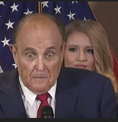 Rudy Giuliani, looking skeletal and ill, with Jenna Ellis peering auspiciously over his shoulder